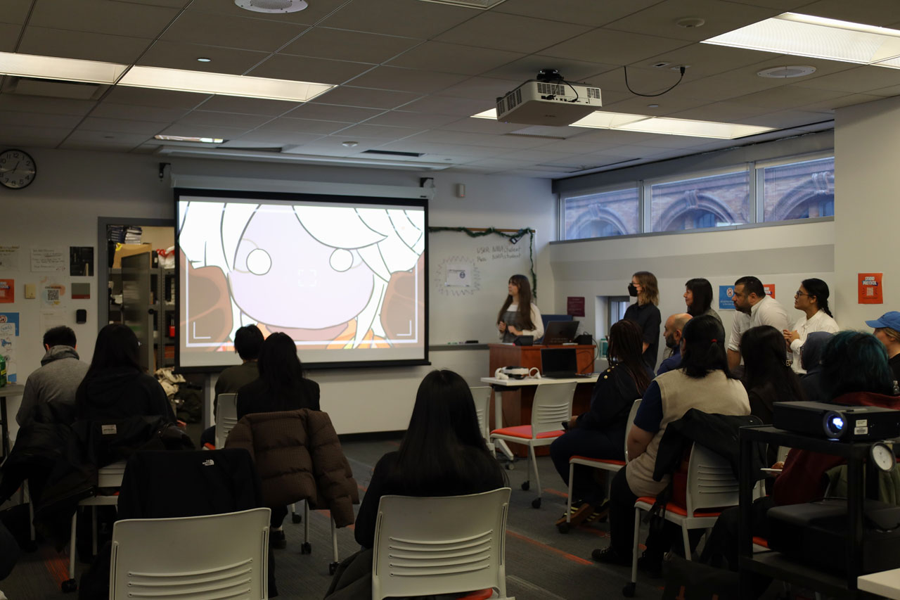 Visitors shown from the behind sitting in rows of orange rolling chairs watching a video screened with a projector in the NMA Studio.