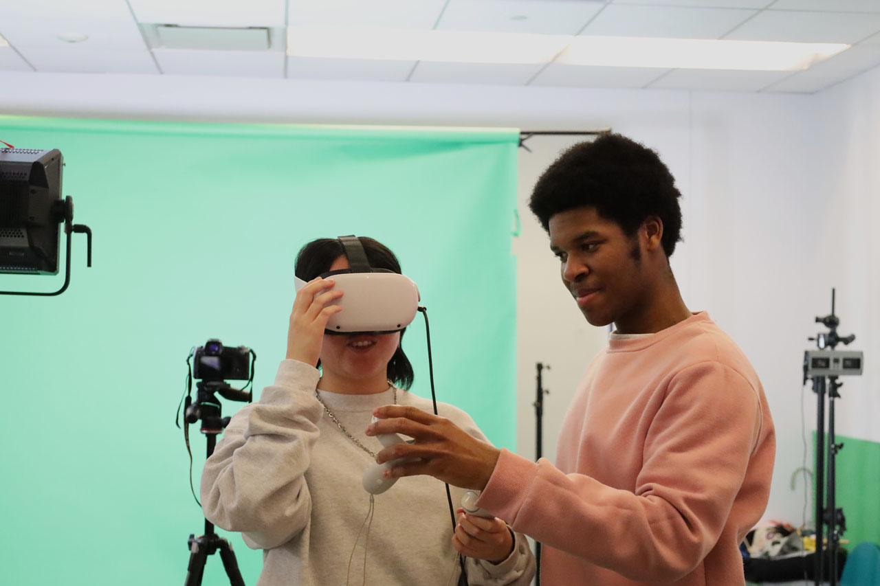 One student tries out a VR headset while another student hands them controllers.