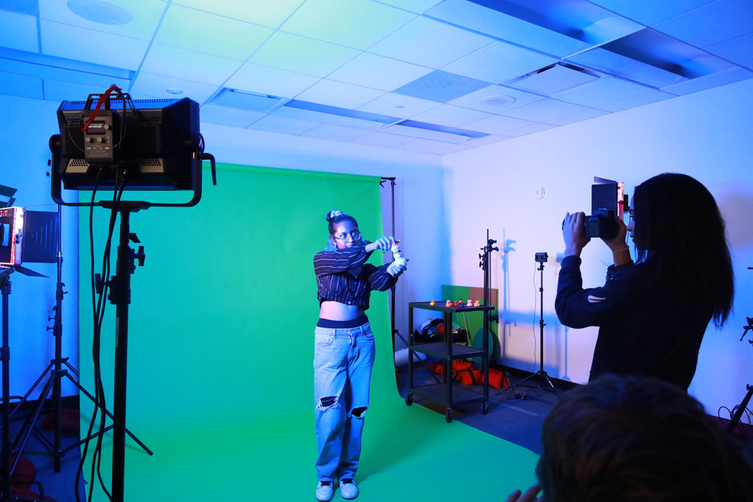 A student posing with a snowman figurine in front of a green screen while another student takes her photo.