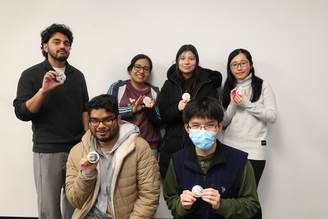 Six smiling students posing with their handmade buttons, four in the back and two crouching in the front.