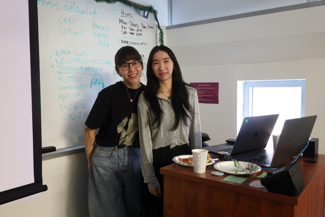 Two docents smiling at the camera, standing by a podium with a laptop on it and front of a whiteboard with notes such as the NMA Studio open hours.