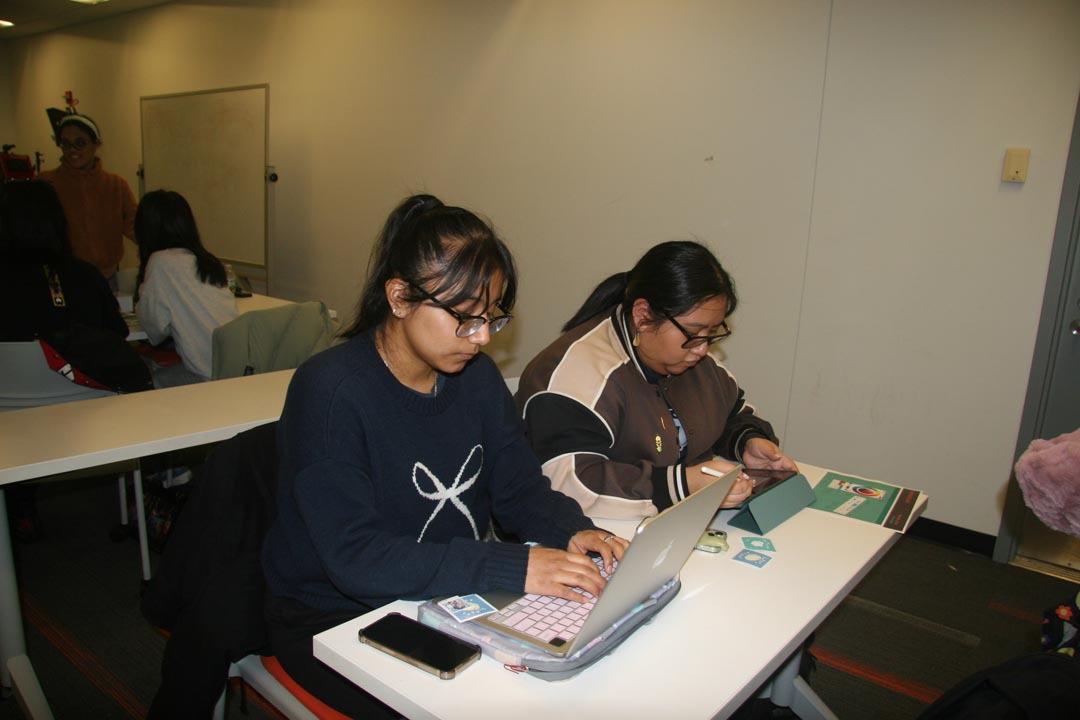 Two students using a laptop and iPad with more students visible in the background.
