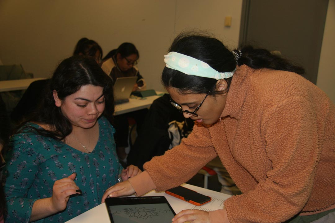 A person with dark hair and a white headband leaning over to help a person with dark long hair use Procreate on an iPad. The iPad shows a line illustration of a flower.
