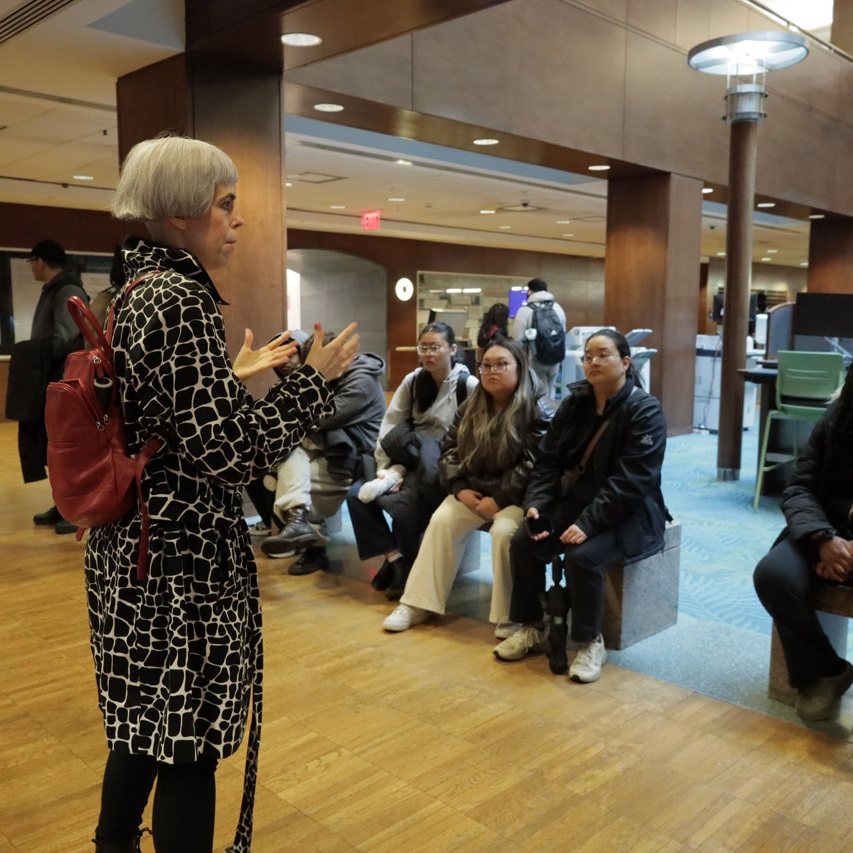 Colleen Asper, a white woman with a bleached short, thick bob wearing a long black and white leopard-print coat, speaks to a group of students.
