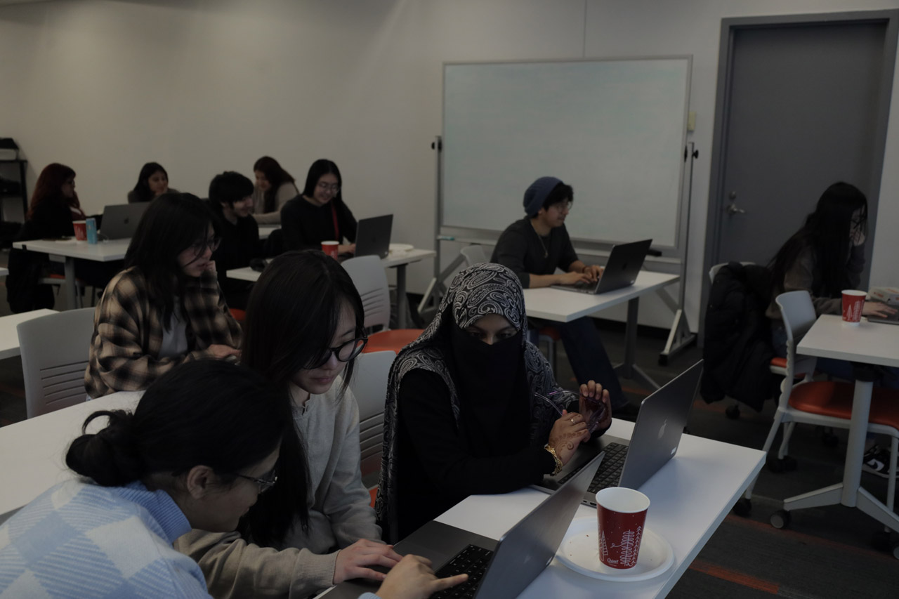 Students working on laptops at rectangular tables. A docent leans over one student, providing one-on-one support.