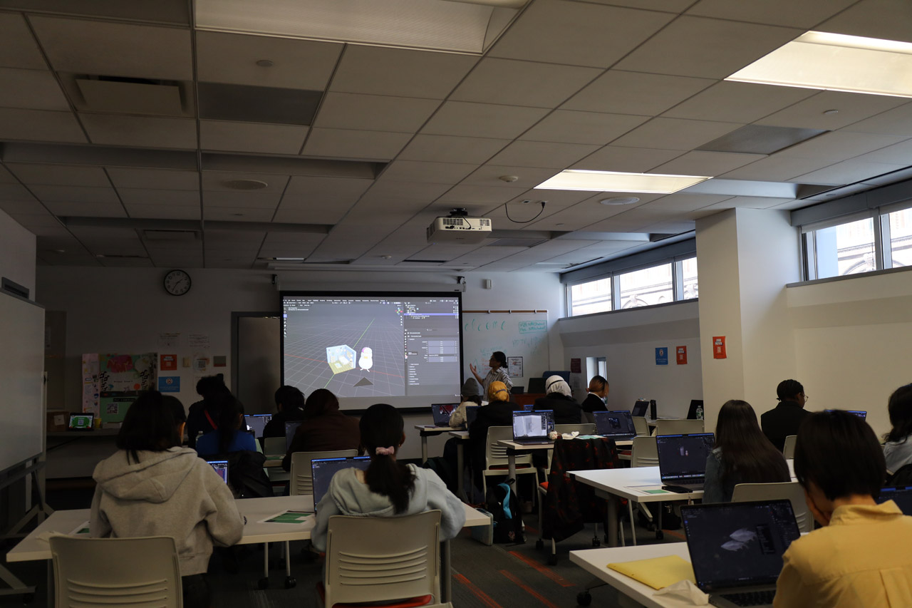 Students sitting at tables in the NMA Studio with a docent at a podium in the front of the room. Blender is open on the projector screen.