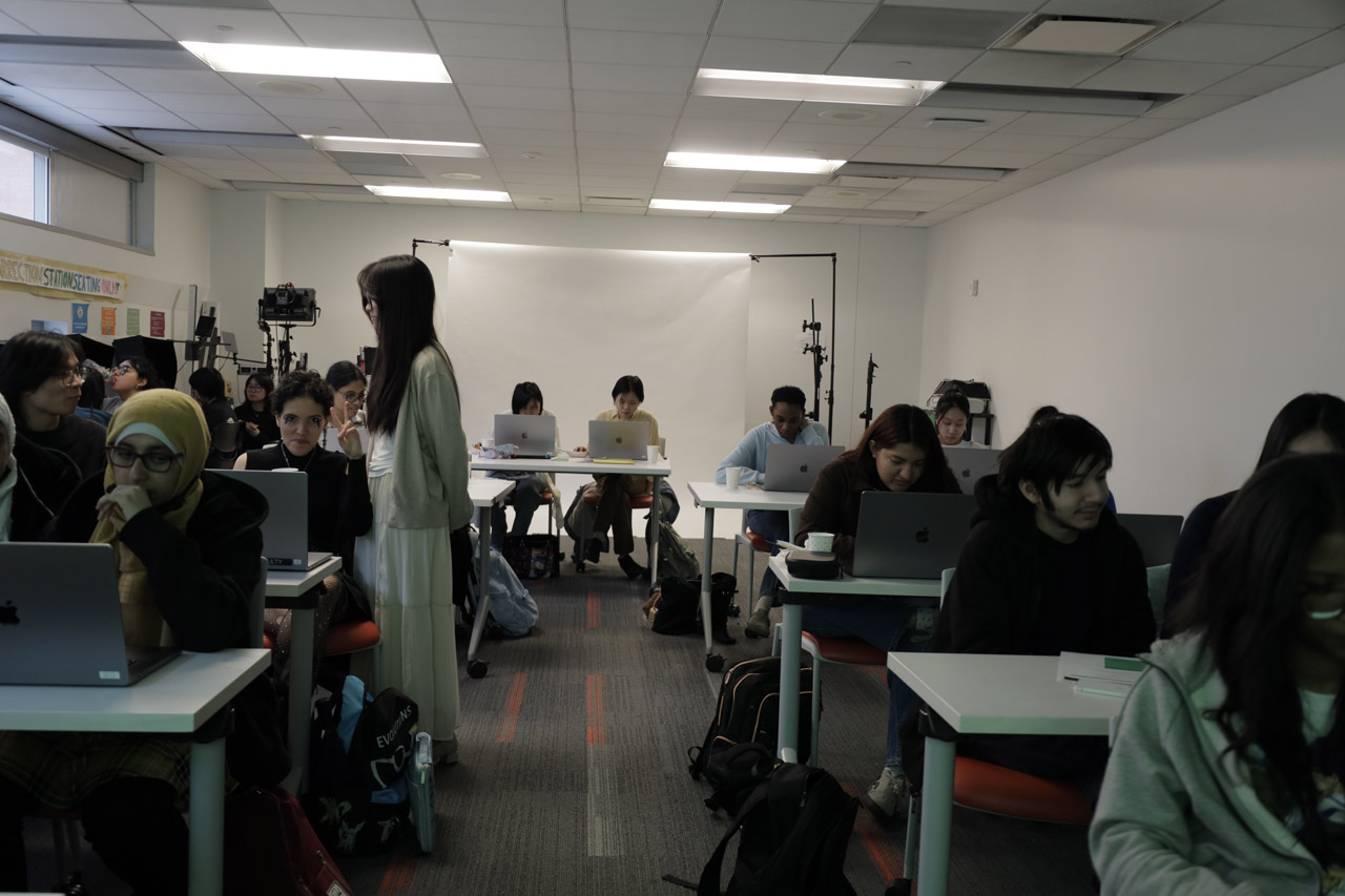 Students working on laptops at rectangular tables. One makes a peace sign at the camera.