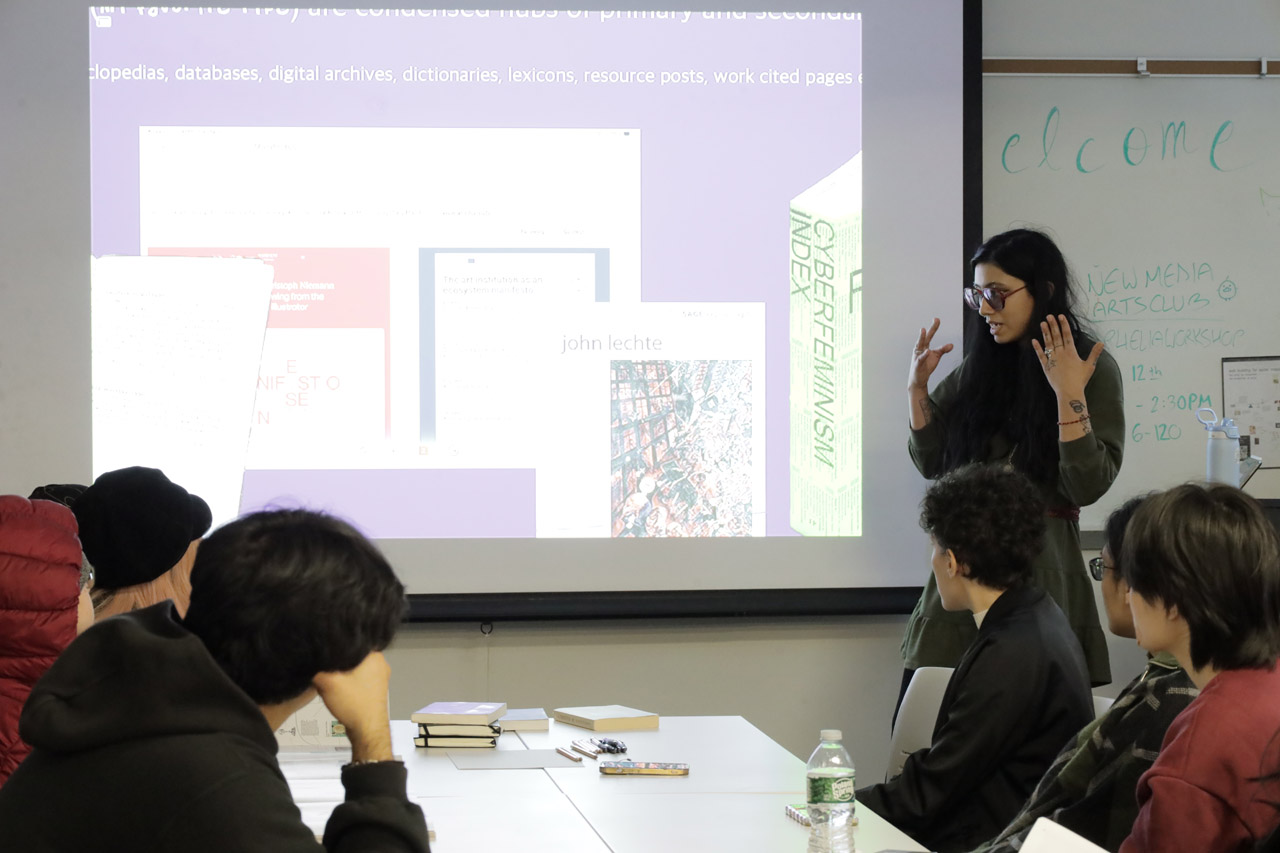 A woman with long black hair presents about primary and secondary sources. Books lay on a table with students seated around it.